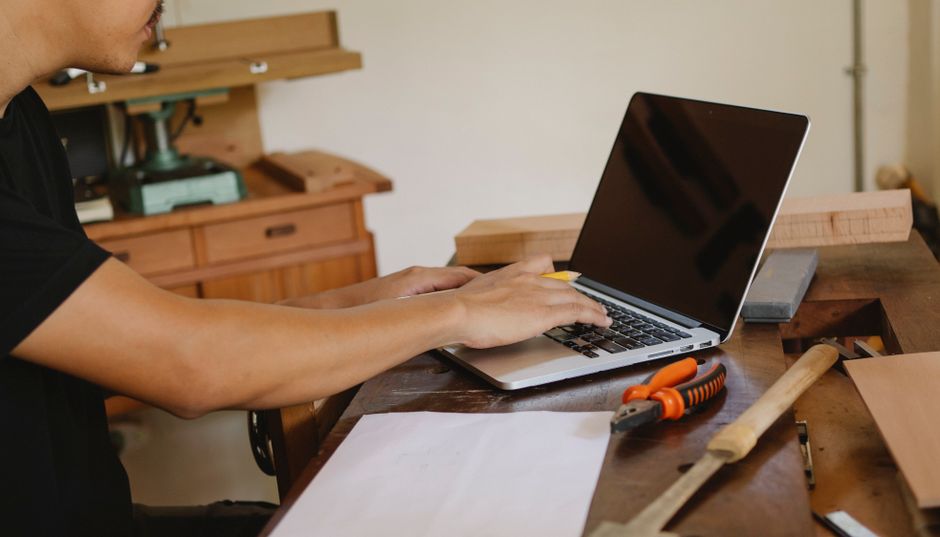 Pessoa utilizando um laptop em um ambiente de trabalho artesanal ou de marcenaria, com ferramentas e materiais de madeira ao redor.