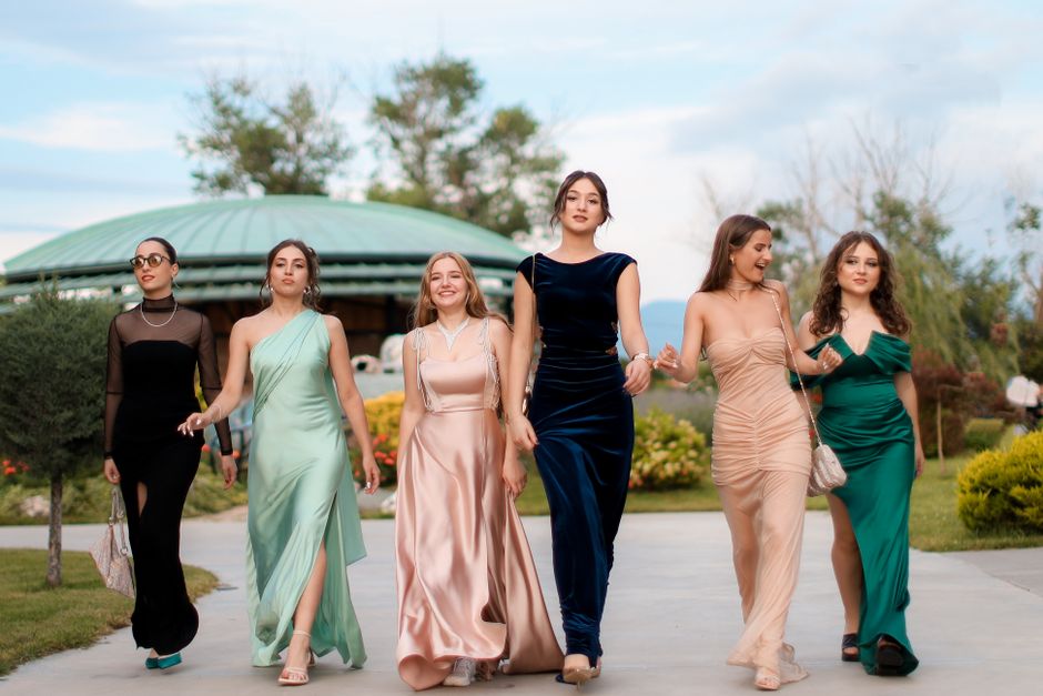 Grupo de mulheres elegantes caminhando ao ar livre em um evento social ou festa, vestidos sofisticados e ambiente natural ao fundo.