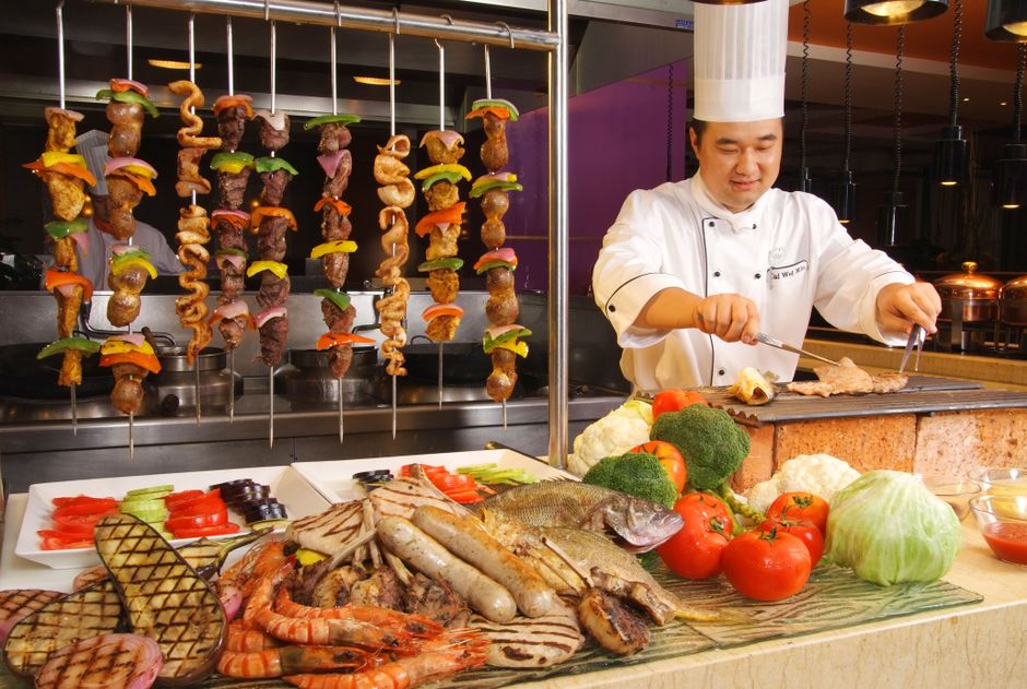 Chef preparando carne e legumes em uma estação de churrasco com vegetais frescos e espetos de carne ao fundo, em um ambiente de gastronomia variada.
