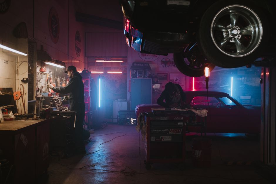 Imagem de uma oficina de carros com iluminação neon, onde duas pessoas trabalham em diferentes tarefas sob um ambiente industrial e moderno.