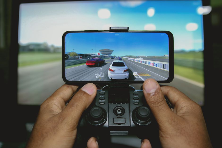 Pessoa jogando um jogo de corrida em um celular conectado a um controle, em frente a uma tela grande de vídeo game ou monitor.