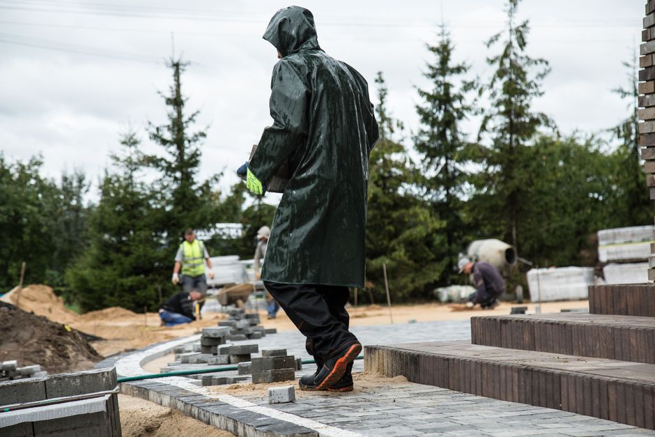 Pessoa trabalhando na construção de um passeio com conduíte na área externa sob clima chuvoso, com outros operários ao fundo.