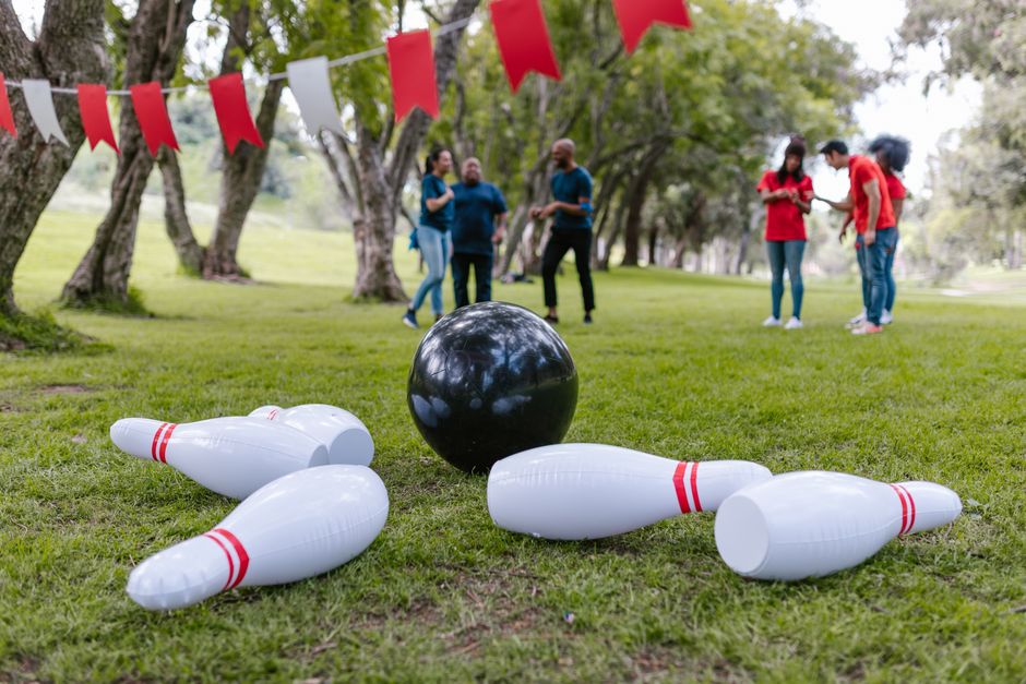 Pessoas em um evento ao ar livre se divertindo com uma atividade de boliche inflável sob árvores, com decoração de bandeirinhas coloridas.