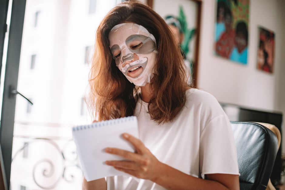 Mulher com máscara facial, lendo ou analisando um caderno em um ambiente de escritório ou estudo, transmitindo sensação de autocuidado ou rotina de bem-estar.