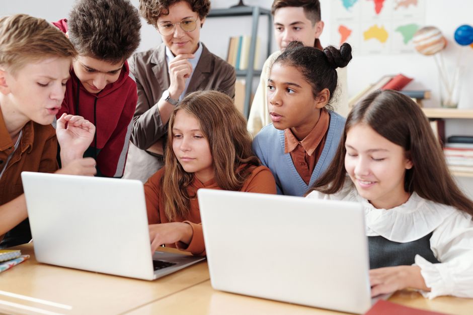 Grupo de estudantes e professora reunidos ao redor de laptops durante uma atividade de aprendizagem colaborativa na sala de aula.