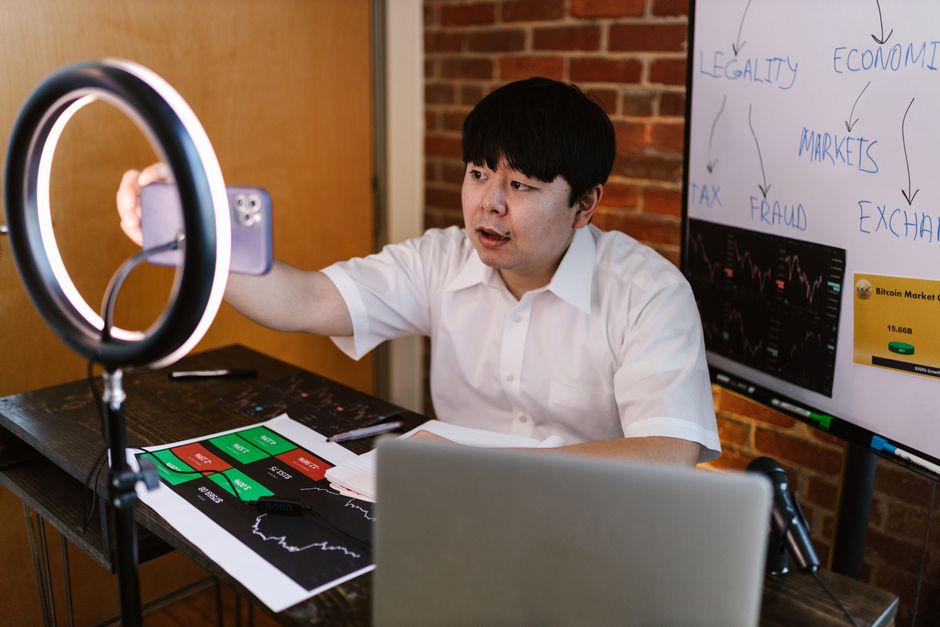 Homem gravando vídeo para blog sobre análise de mercado financeiro, com computador, gráficos e quadro de anotações ao fundo.