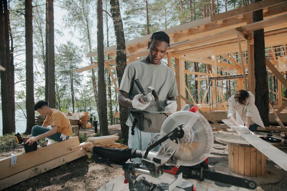 Homem trabalhando na construção de uma casa de madeira em meio a uma floresta, com outros colegas ao fundo realizando tarefas relacionadas à obra.