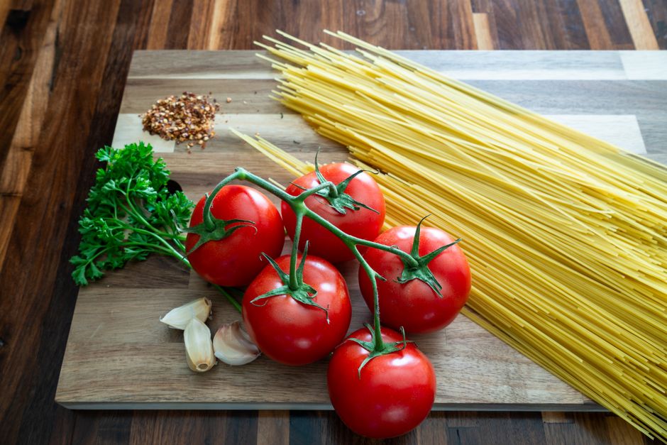 Imagem de ingredientes frescos de culinária italiana, destacando tomates, alho, salsinha, pimenta do reino e macarrão cru, prontos para preparo de uma receita.