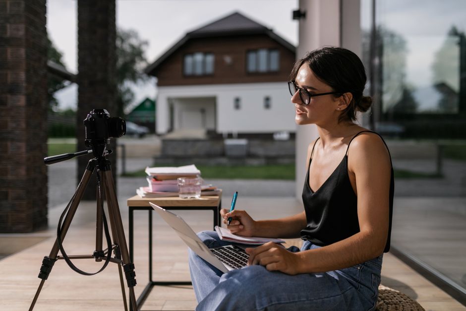 Mulher estudando ou criando conteúdo ao ar livre com câmera, laptop e materiais de escrita, em um ambiente tranquilo e moderno.