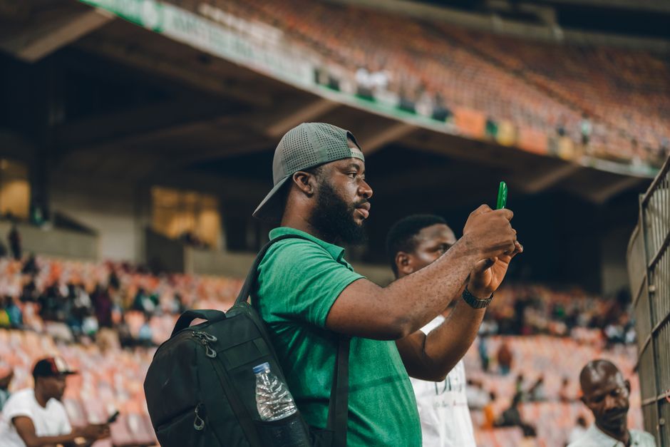 Homem assistindo a um evento esportivo, registrando o momento com o celular no estádio, destacando o envolvimento e a paixão pelo esporte.