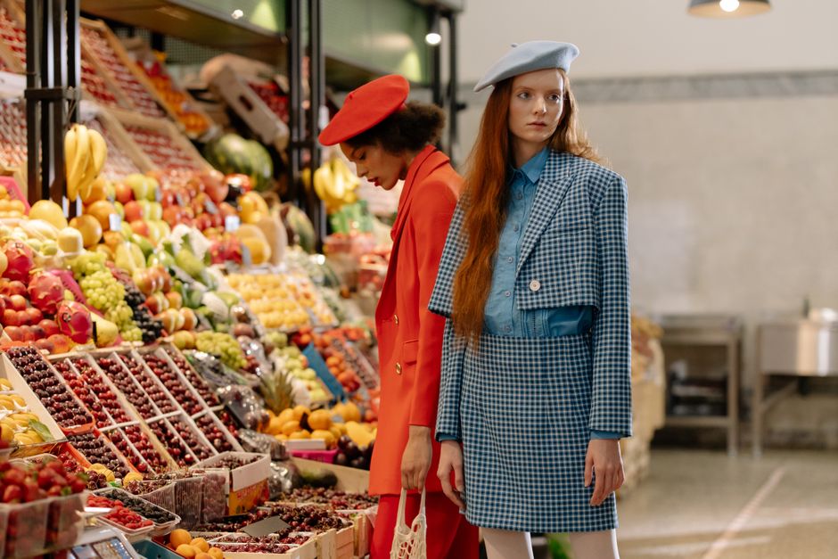 Duas mulheres em estilo fashion, usando roupas coloridas vintage, explorando uma seção de frutas em uma loja ou mercado, com uma atmosfera moderna e descontraída.