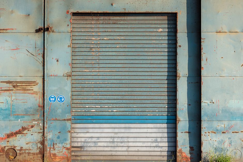 Imagem de uma porta de metal antiga e enferrujada em uma parede desbotada, transmitindo sensação de abandono ou resistência.