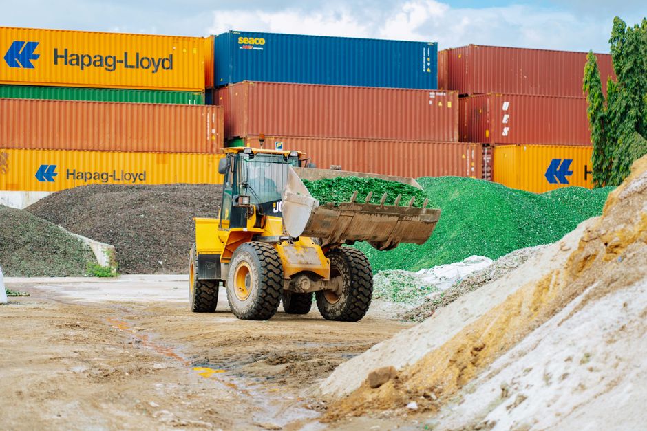 Máquina de construção movendo material em um cenário logístico com contêineres ao fundo, ilustrando transporte e armazenamento de cargas.