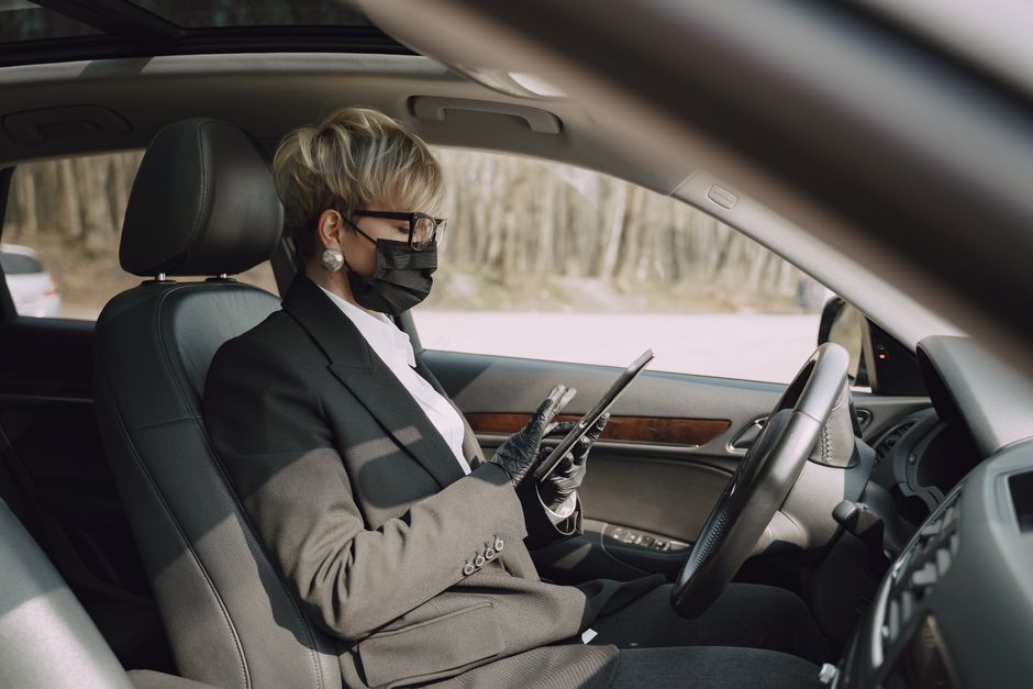 Mulher de terno e máscara usando um tablet enquanto dirige um carro, simbolizando trabalho remoto ou teletrabalho em viagens.