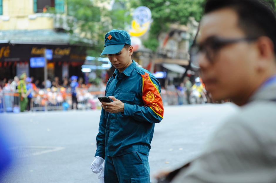 Imagens de um jovem policial militar verificando informações pelo celular durante uma operação na rua, com pessoas ao fundo, em um ambiente urbano em movimento.