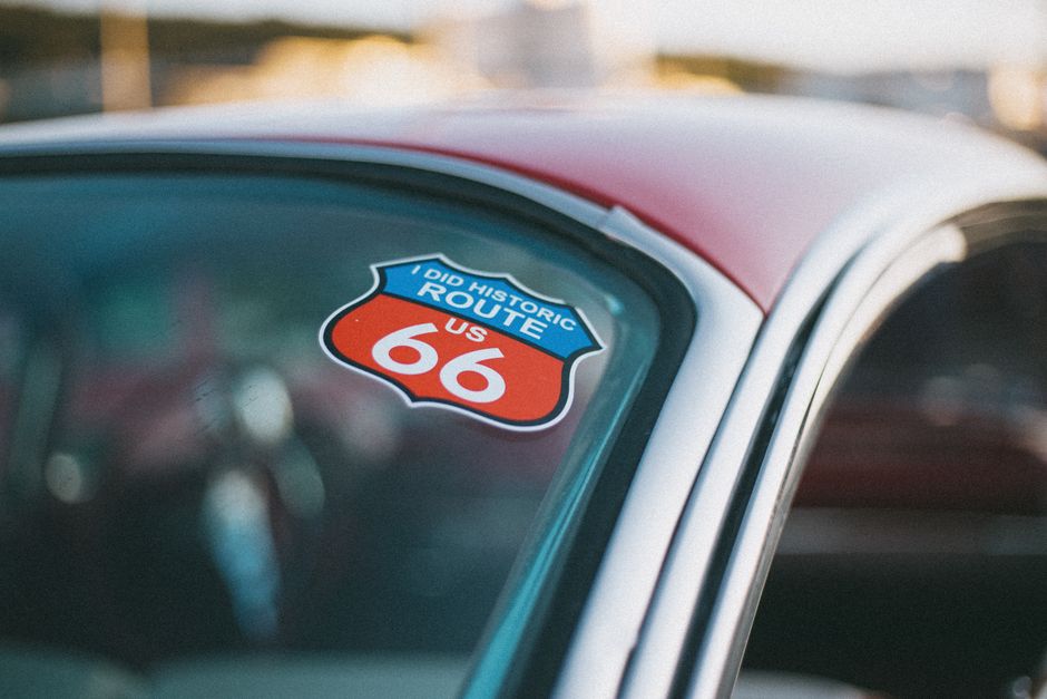 Imagem de um carro antigo com uma foto de um ônibus nas costas e um adesivo de rota histórica da U.S. Route 66 na janela dianteira.