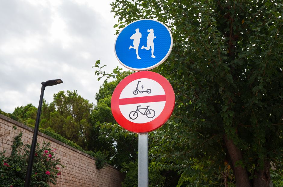 Placa de trânsito indicando áreas para pedestres e restrição para ciclistas, em um ambiente urbano com árvores e iluminação pública ao fundo.