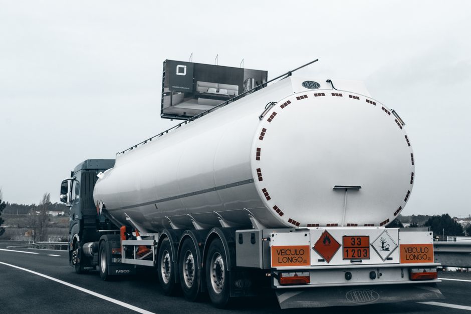 Imagem de um caminhão-tanque transportando líquidos inflamáveis, exibindo placas de risco, em uma rodovia com céu nublado.