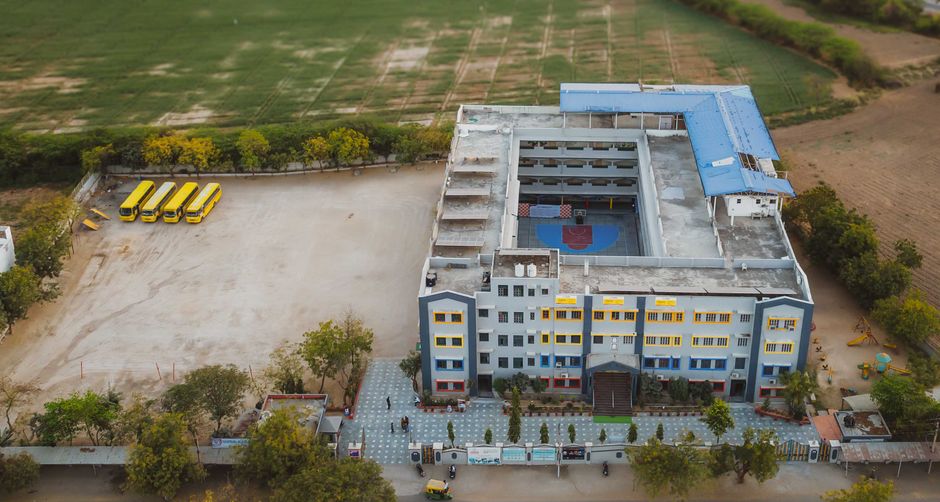 Imagem aérea de uma escola com quadra poliesportiva e área de lazer, localizada em uma área rural ou semi-rural.