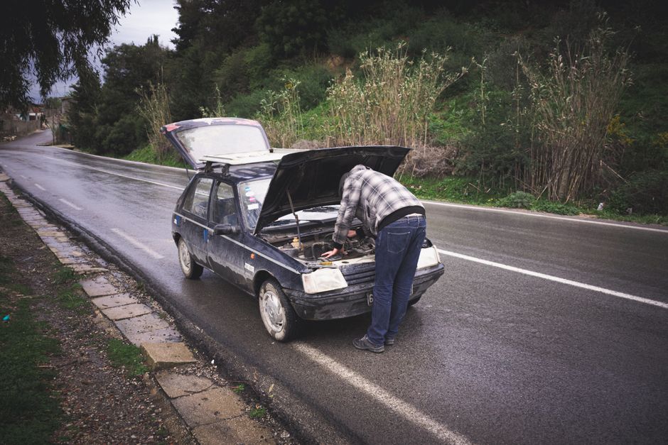 Pessoa realizando manutenção ou inspeção em um carro parado na rua, com o capô levantado e o motor aberto.