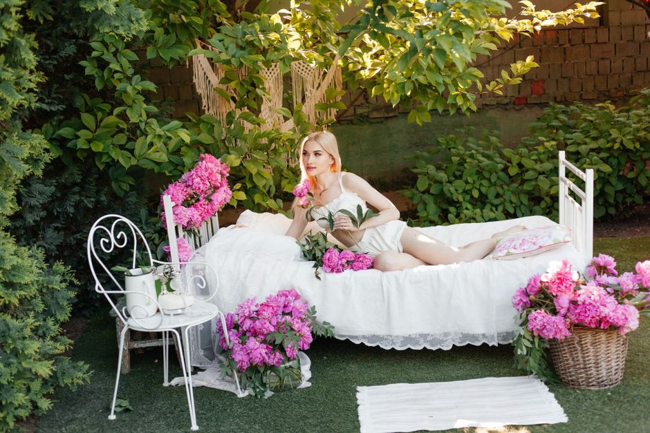 Mulher posando deitada em uma cama ao ar livre, cercada por plantas e flores, em um ambiente de jardim.