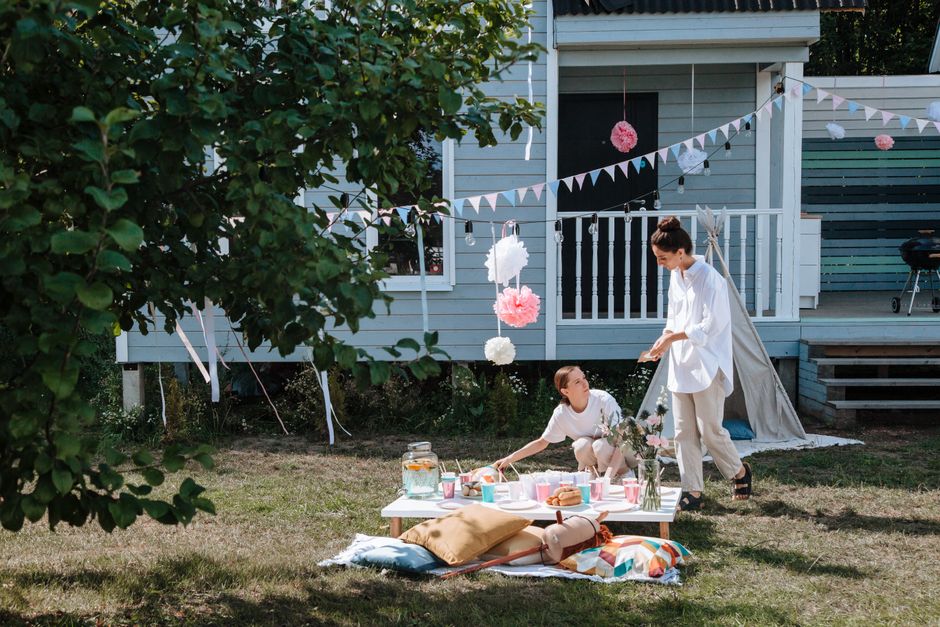 Duas mulheres organizando um piquenique ao ar livre em um espaço com decoração festiva, em um jardim próximo a uma casa de madeira.