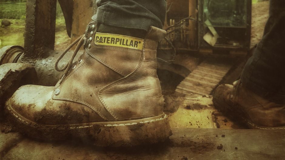 Imagem de botas de trabalho de couro em um ambiente de área rural ou de construção, destacando o esforço físico e a dedicação ao trabalho ao ar livre.