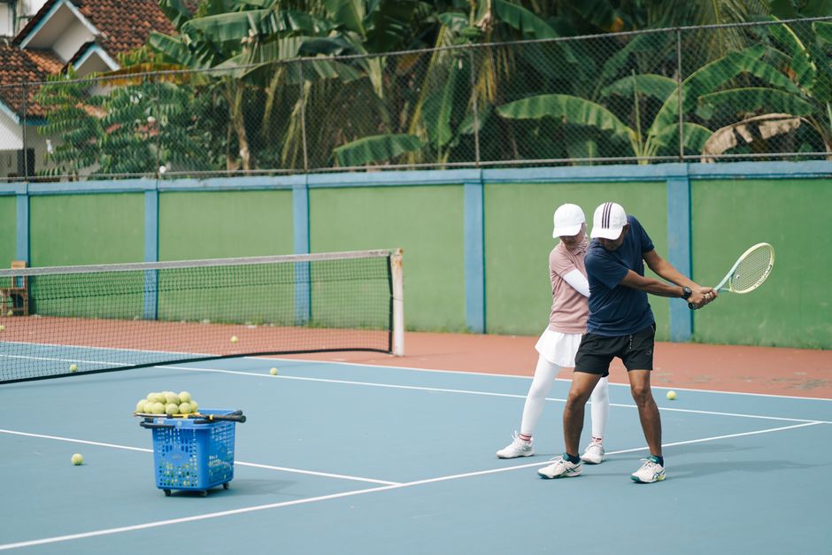 Dupla praticando tênis em uma quadra ao ar livre, enfatizando o aprendizado e a prática esportiva para iniciantes.