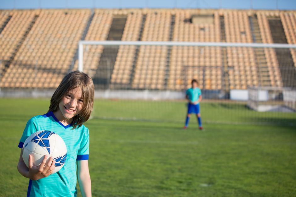 Menina sorridente segurando uma bola de futebol em um campo de estádio, com uma goleira ao fundo treinando.