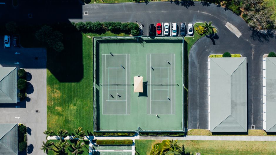Imagem aérea de uma quadra de tênis com pessoas jogando, em um ambiente de lazer ao ar livre, ao lado de uma área residencial ou de condomínio.