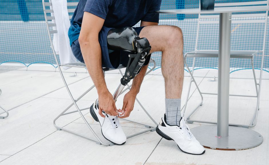 Homem com prótese na perna ajustando o tênis na varanda, evidenciando recuperação e adaptação à nova tecnologia.
