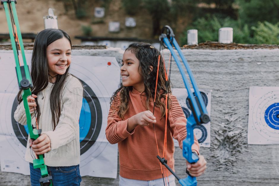 Duas crianças brincando de arco e flecha ao ar livre, simbolizando diversão e aprendizado na prática de esportes ao ar livre.
