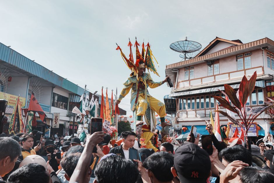 Pessoa participando de uma feira cultural ou desfile tradicional com elementos coloridos e figurinos vibrantes, em ambiente urbano ao ar livre.