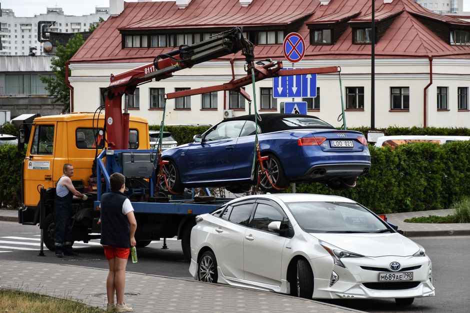 Imagem de um guincho elevando um carro conversível azul para remoção de um veículo estacionado irregularmente, com uma pessoa observando e outro pendurado na traseira do caminhão.