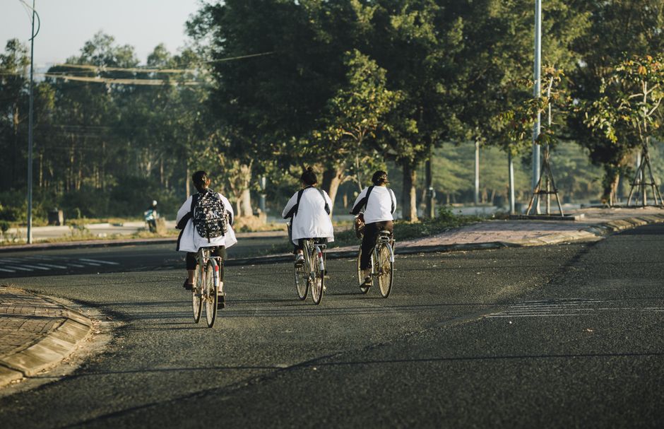 Imagem de estudantes seguindo de bicicleta por uma rua arborizada durante o dia, simbolizando movimento e sustentabilidade no contexto escolar.