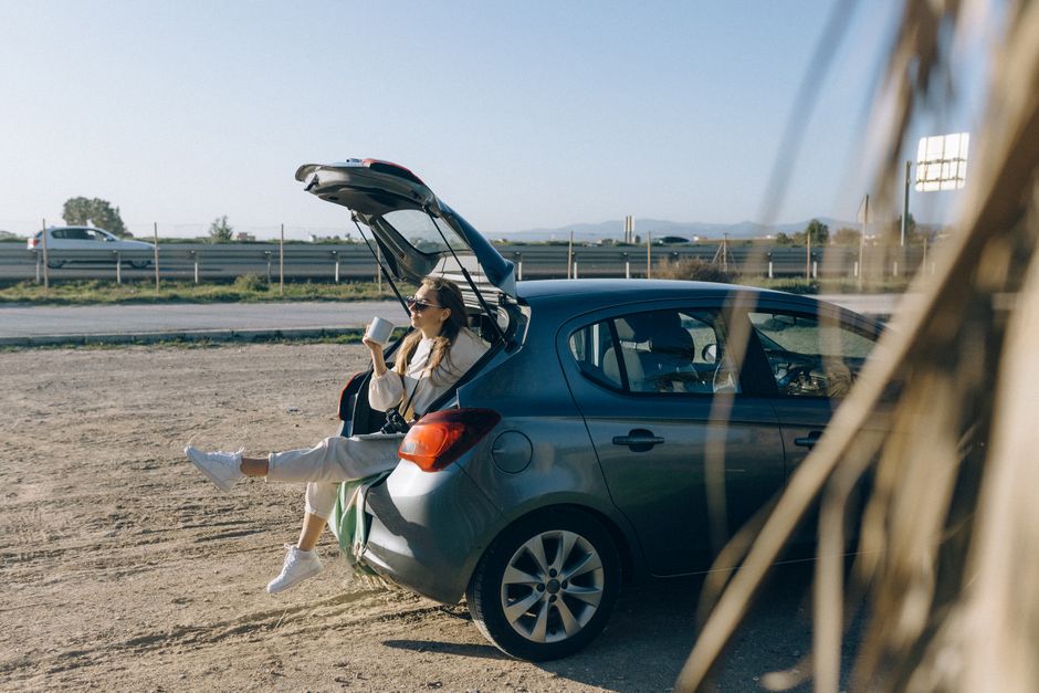 Mulher relaxando no porta-malas de um carro ao ar livre, tomando café em um cenário de viagem ou aventura.