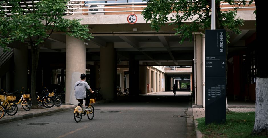 Homem pedalando por uma área de acesso de edifício universitário, destacando o ambiente acadêmico e mobilidade urbana.