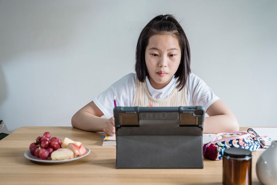 Jovem estudante assistindo aula online com atenção em seu tablet, com lanche e materiais de estudo ao lado, em ambiente doméstico.