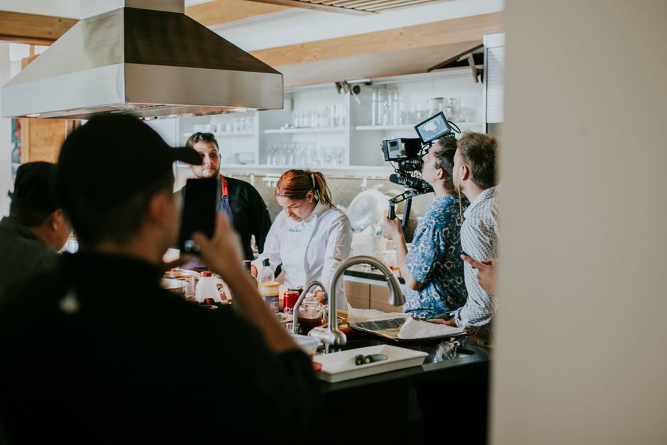 Cena de gravação de um programa de culinária em uma cozinha moderna, com equipe de filmagem e chef preparando alimentos.