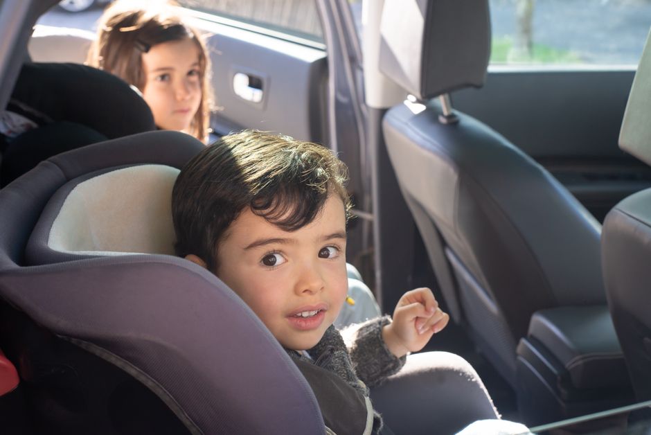 Crianças sentadas em assentos de carro durante uma viagem, com uma delas olhando para a câmera.
