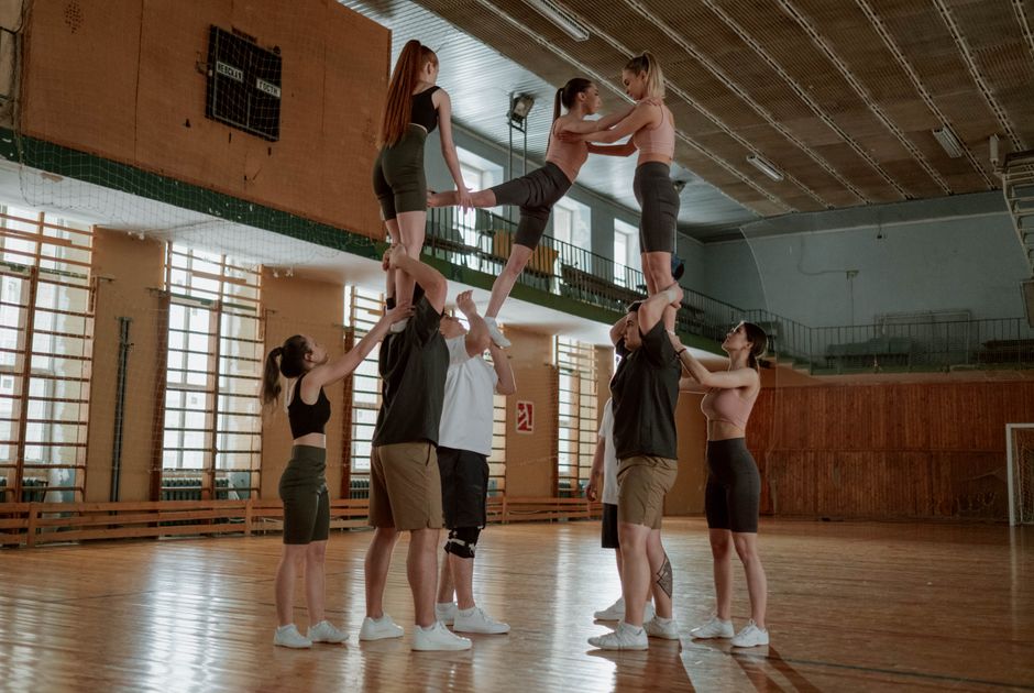 Grupo de jovens praticando ginástica acrobática em ginásio com estrutura de madeira e janelas amplas ao fundo.