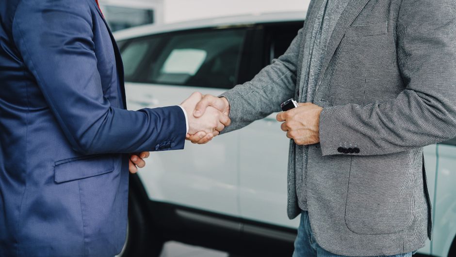 Imagem de dois homens de negócios apertando as mãos em sinal de acordo ou parceria ao lado de um carro, representando negócios ou negociações comerciais.