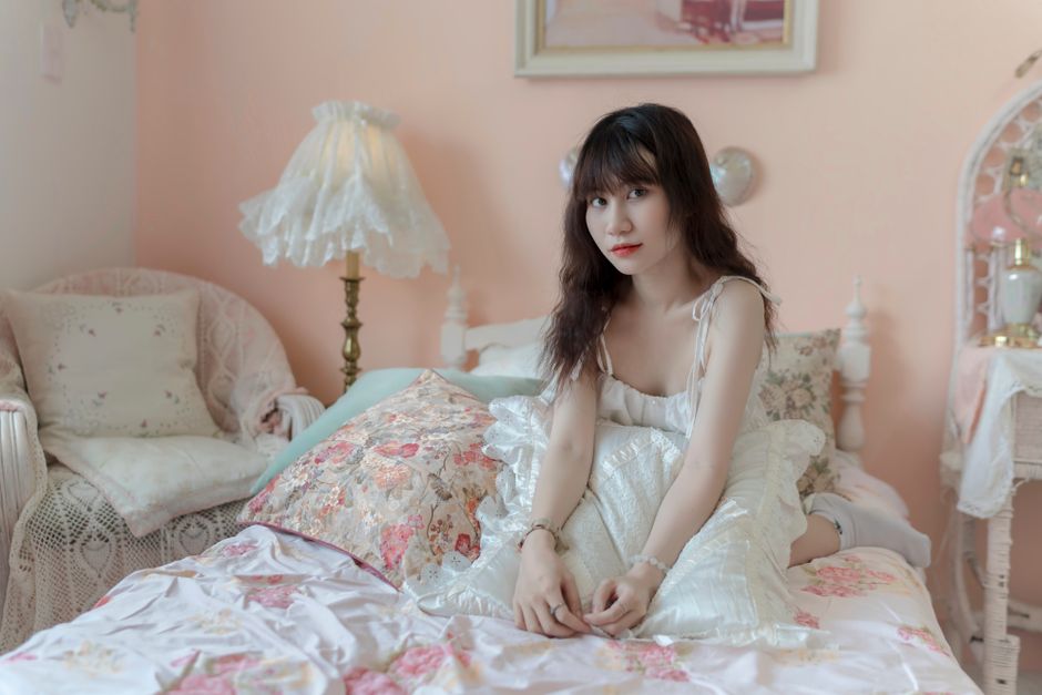Mulher jovem com expressão pensativa, sentada de frente para a câmera em quarto decorado com estilo romântico, com móveis e objetos delicados em tons pastel.