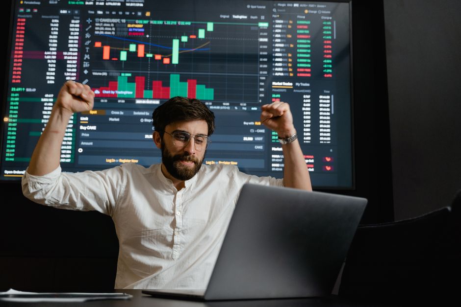 Homem comemorando vitória financeira diante de tela com gráficos de mercado e ações, simbolizando sucesso em investimentos.