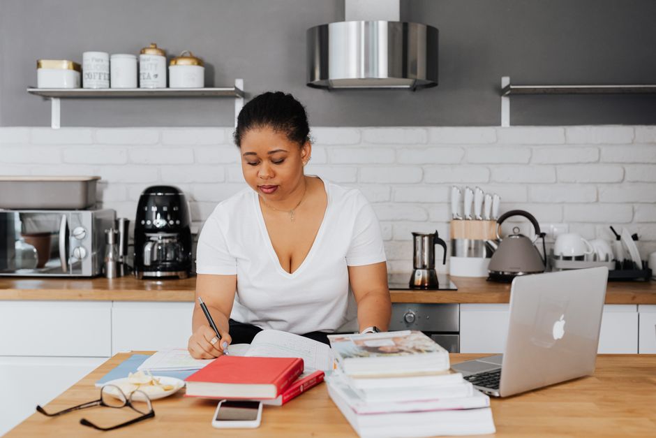 Mulher estudando ou trabalhando em casa com materiais de estudo e tecnologia, em ambiente de cozinha moderna.