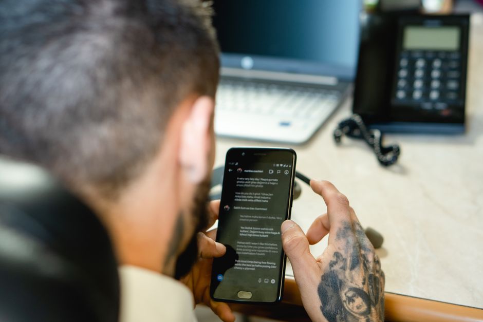 Pessoa usando smartphone em ambiente de trabalho, com laptop e telefone fixo ao fundo, ilustrando comunicação corporativa ou rotina profissional remota.