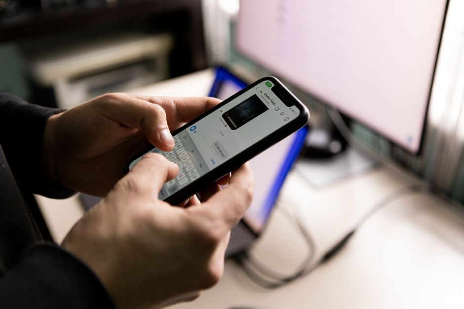 Pessoa utilizando um smartphone para acessar informações ou fazer tarefas relacionadas a negócios ou finanças, com computadores ao fundo.