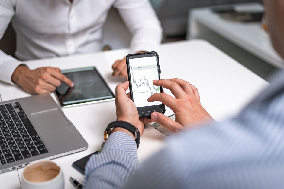 Duas pessoas analisando dados financeiros em uma reunião de negócios, utilizando dispositivos digitais e gráficos de desempenho.