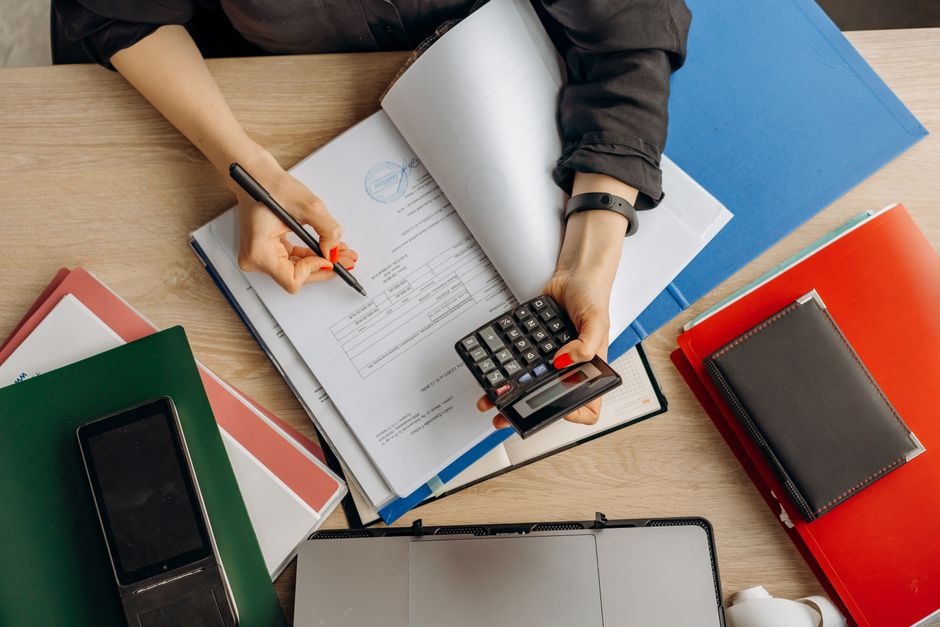 Pessoa analisando e preenchendo documentos financeiros em um ambiente de escritório, com calculadora e materiais de papel ao redor.