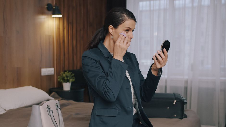 Mulher se arrumando no quarto enquanto se prepara para sair, usando maquiagem e conferindo o espelho, transmitindo uma rotina de cuidados pessoais e preparação para um compromisso.